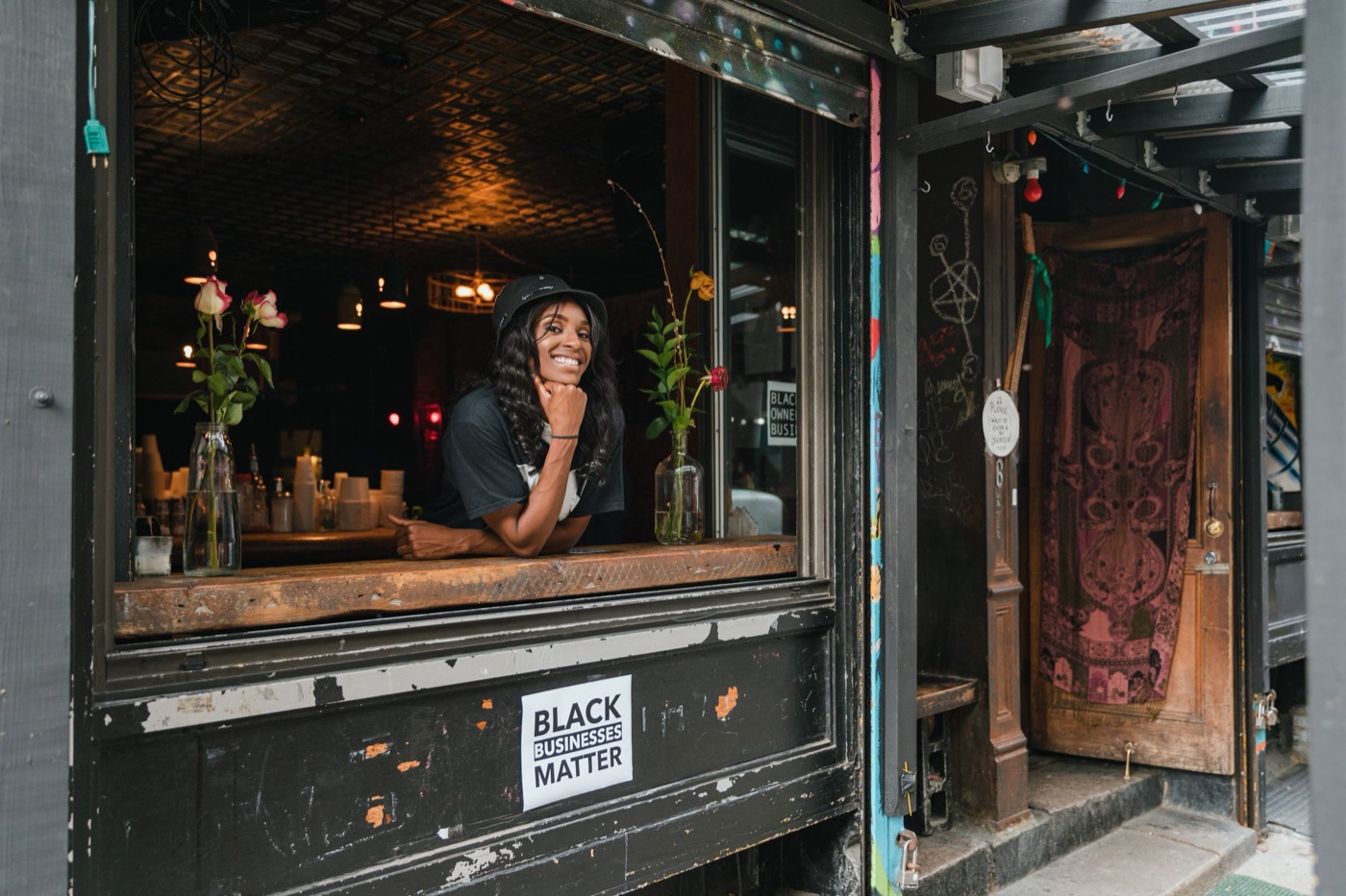 A smiling Black woman leans out of the open front window of her cafe, with flowers on the sill and a Black Businesses Matter sticker below
