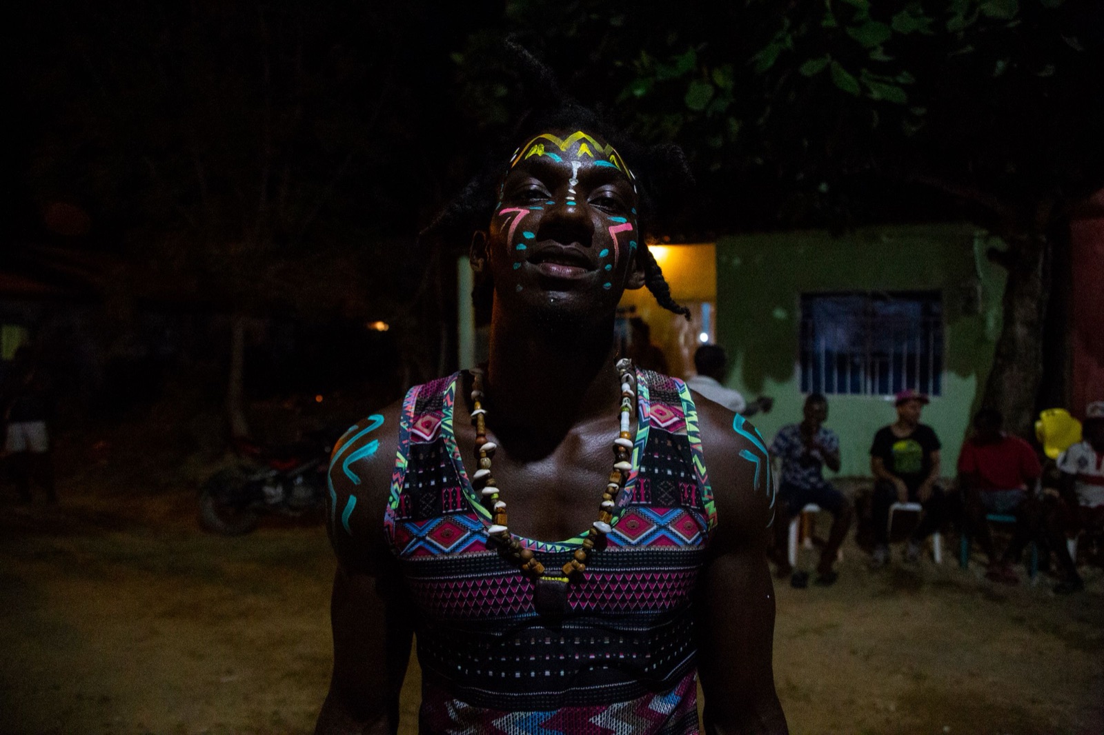 A young man from San Basilio de Palenque in festival face paint, beads and a patterned top, with neighbors gathered behind him at night