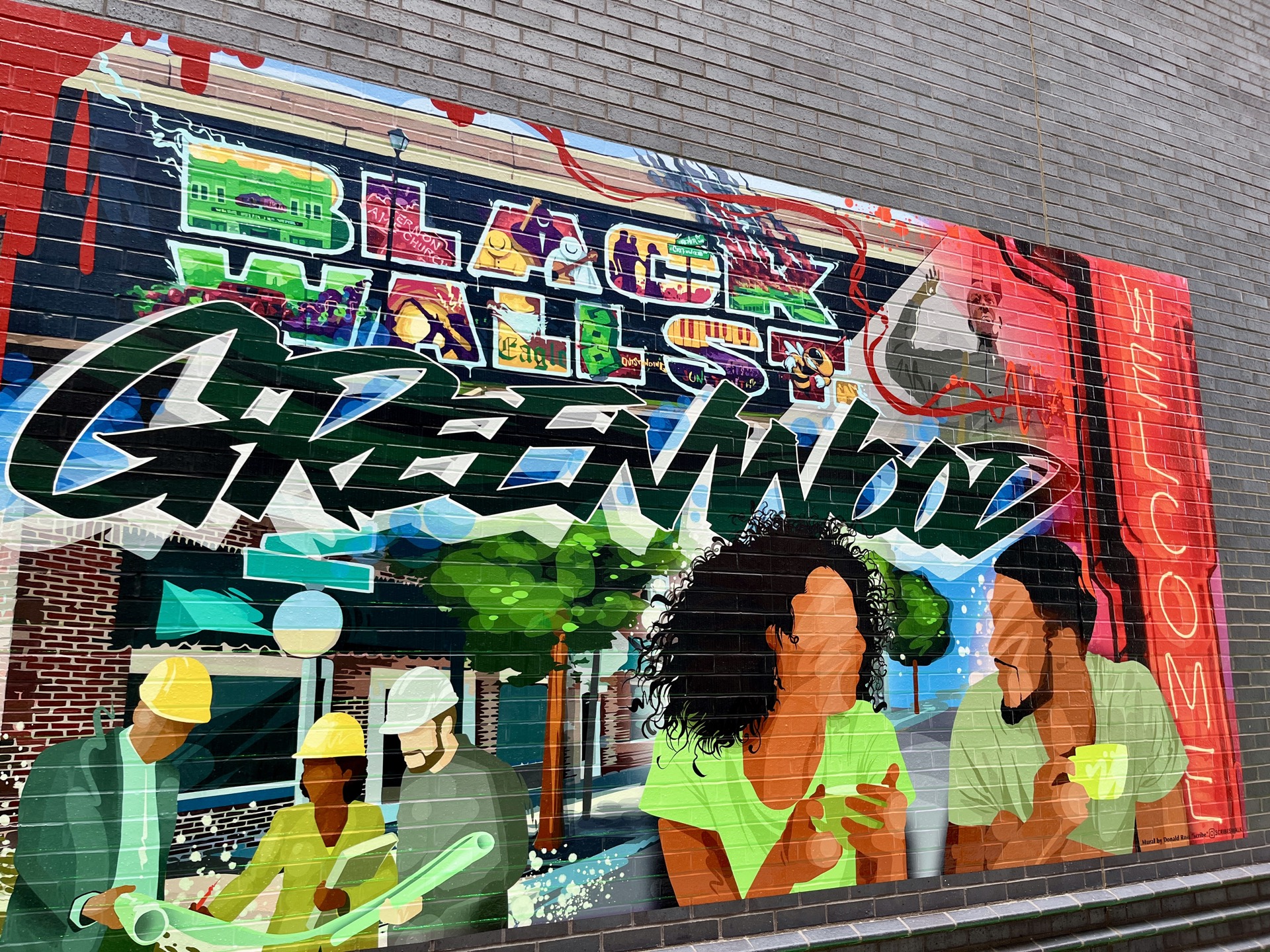 A mural on a brick wall in Greenwood reading Black Wall Street and Greenwood, with painted figures of builders in hard hats and neighbors gathered on the avenue