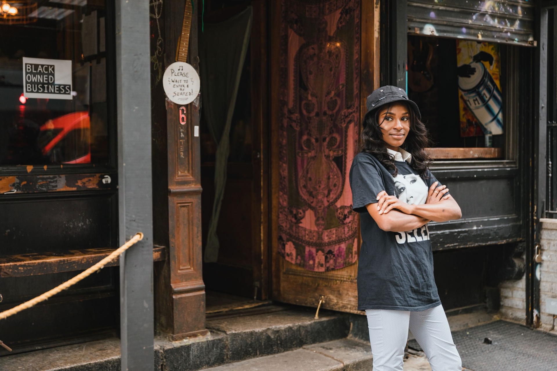A smiling Black woman in a cap and T-shirt stands with her arms crossed outside a storefront with a Black Owned Business sign in the window